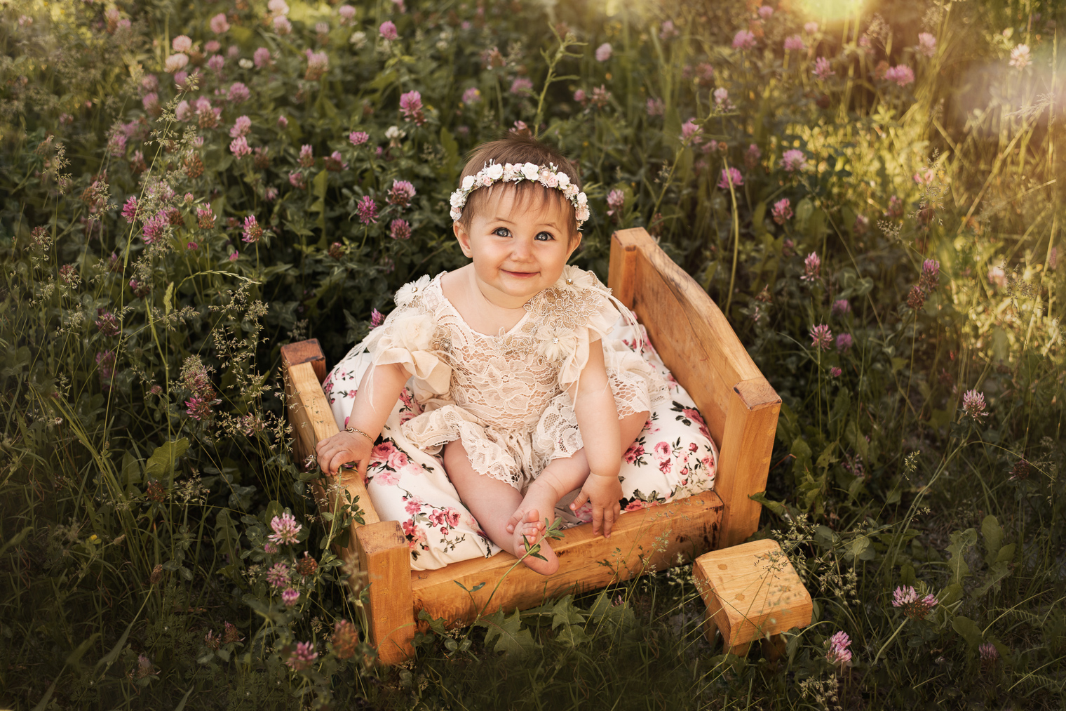 Bébé fille qui sourit, elle a une robe en dentelle, une couronne de fleurs et elle est assise sur un petit lit en bois au milieu d'un champ de fleurs de trèfle