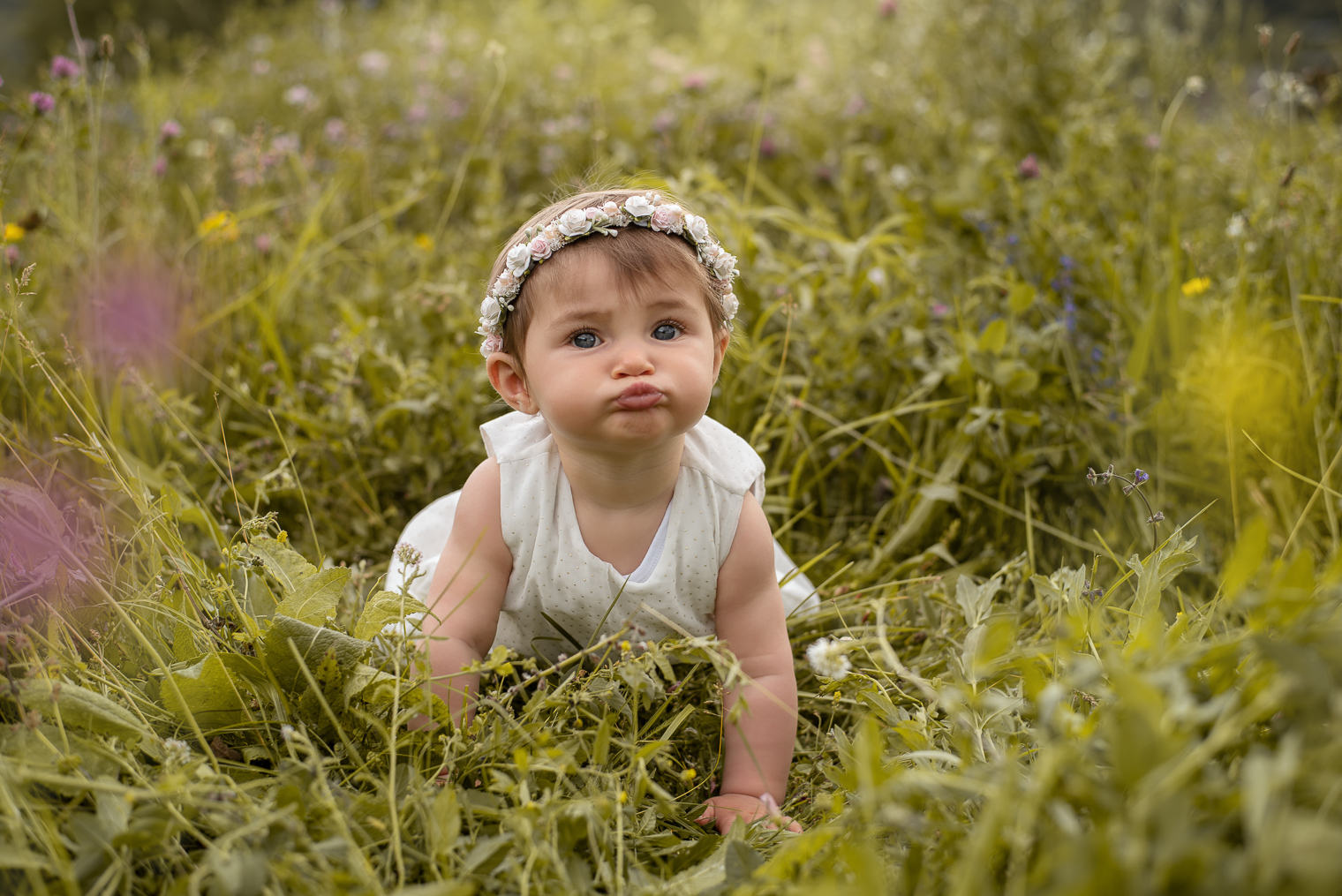 Bébé fille avec une robe blanche a pois doré et une couronne de fleurs, elle est à quatre patte dans l'herbe et fait une grimace