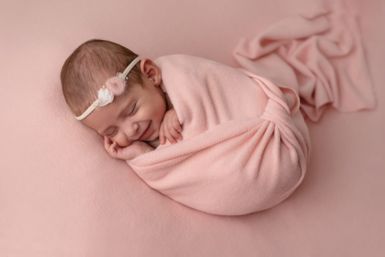 Bébé fille qui sourit, emmaillotée dans un tissu rose avec un bandeau rose et blanc