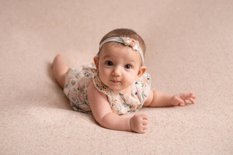 Bébé fille avec une robe à fleurs et un bandeau assorti, allongé sur le ventre sur un tissu beige chiné