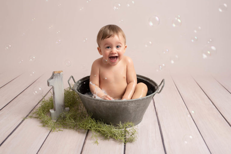 Bébé garçon assis dans une bassine en métal remplie de lait, il a un grand sourire, des bulles de savon volent autour de lui
