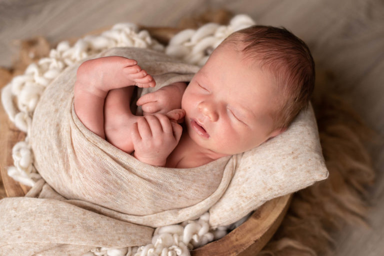 Nouveau-né garçon emmailloté en position foetale dans un tissu chiné beige, il tient son pied dans sa main