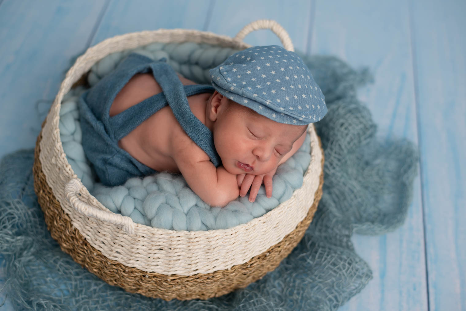 Nouveau-né garçon allongé sur le ventre dans un panier, sur une couverture en laine bleu, il porte une salopette et un béret bleu avec des petites étoiles blanche