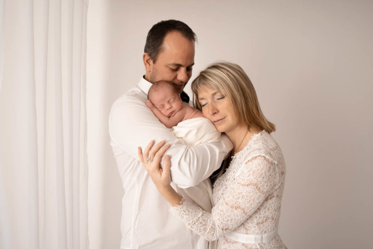 Deux parents et leur bébé, le papa porte le nouveau-né dans ses bras et la maman pose sa tête sur lui, tout le monde a les yeux fermés et profite de l'instant câlin