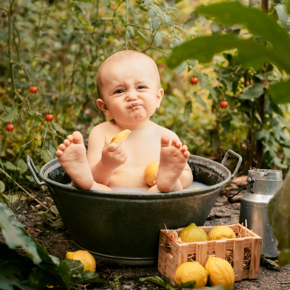 Bébé garçon assis dans une bassine en métal remplie de lait, il goûte une rondelle de citron et fait la grimace