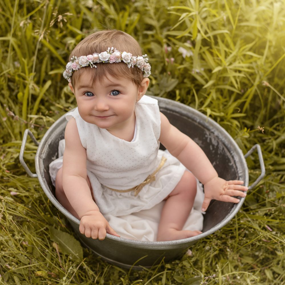 Bébé fille avec une robe blanche a pois doré et une couronne de fleurs, assise dans une bassine en métal dans un champ