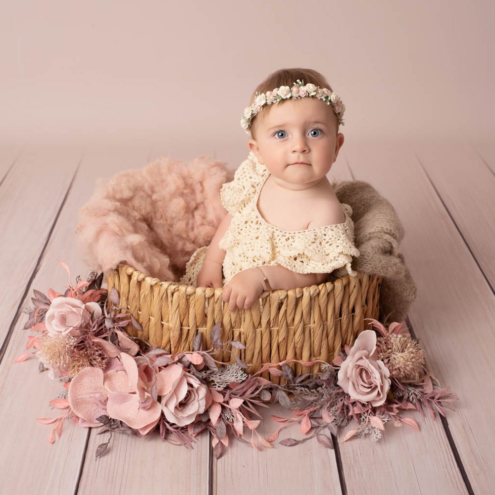 Bébé fille avec une robe en crochet écru et une couronne de fleurs, assises dans un panier en osier avec une couverture en laine rose