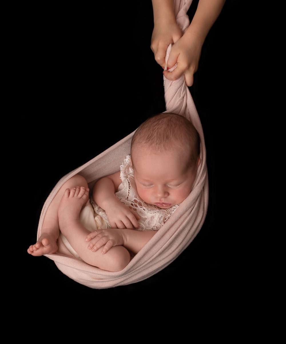 Nouveau-né fille avec un body en dentelle, en position foetale dans un tissu rose pâle, comme suspendu dans les airs et soutenu par les mains de sa grande soeur