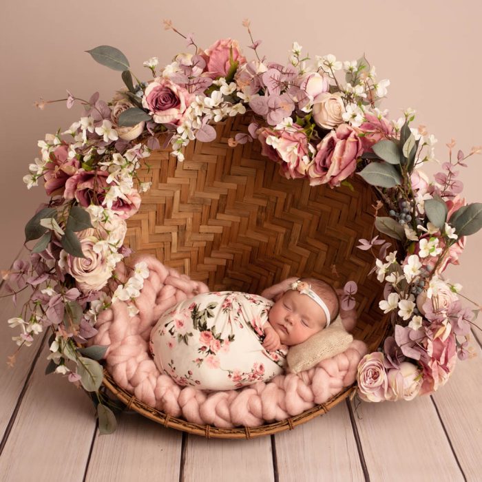 Nouveau-né fille emmaillotée dans une couverture crème avec des fleurs roses, dort dans un abat-jour recouvert d'une guirlande de fleurs
